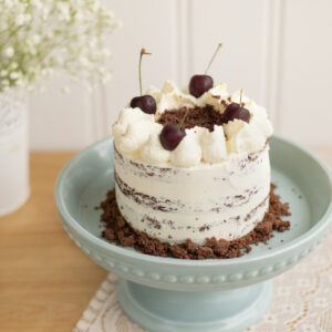 Gâteau d'anniversaire inspiré des saveurs de la Forêt-Noire : gâteau au chocolat, mousse légère à la vanille et cerises. L'extérieur est recouvert d'une fine couche de crème au beurre à la vanille en finition nude, avec une bordure de crumble au cacao et de cerises au sirop en décor. Format 12 parts (7 po de diamètre).