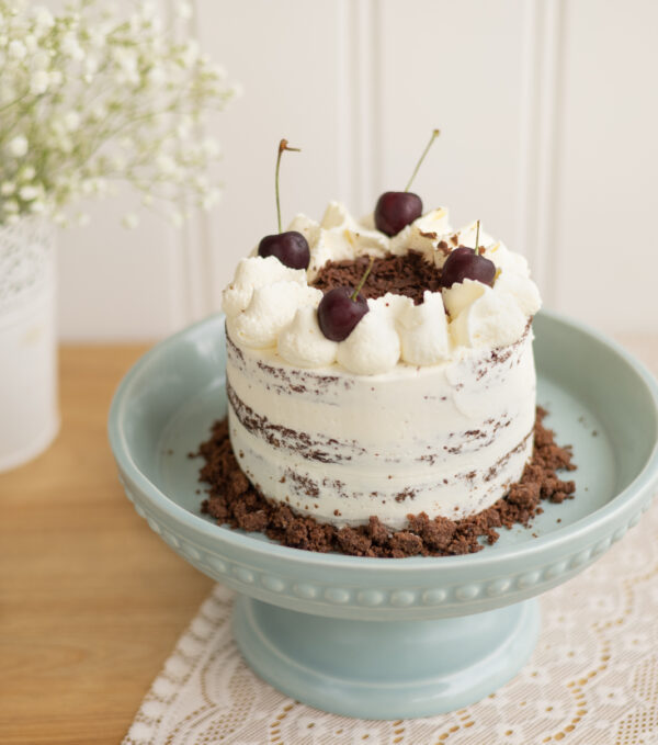 Gâteau d'anniversaire inspiré des saveurs de la Forêt-Noire : gâteau au chocolat, mousse légère à la vanille et cerises. L'extérieur est recouvert d'une fine couche de crème au beurre à la vanille en finition nude, avec une bordure de crumble au cacao et de cerises au sirop en décor. Format 12 parts (7 po de diamètre).